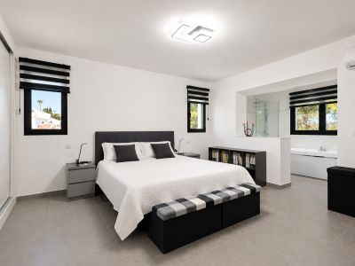 Hedendaagse slaapkamer met geïntegreerde en-suite badkamer, vrijstaand bad en glazen douche. Grijze tegelvloer, witte kasten en uitzicht op groen.