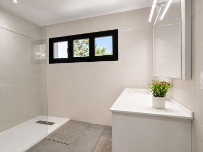 Moderne badkamer met inloopdouche en glazen wand. Lichte tegels, zwart kozijnraam met uitzicht op groen. Wit badmeubel met geïntegreerde wastafel en verlichte spiegel.