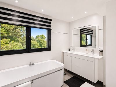 Moderne badkamer met wit vrijstaand bad, dubbele wastafel en zwart kozijnraam met gestreepte jaloezie, uitkijkend op weelderig groen.
