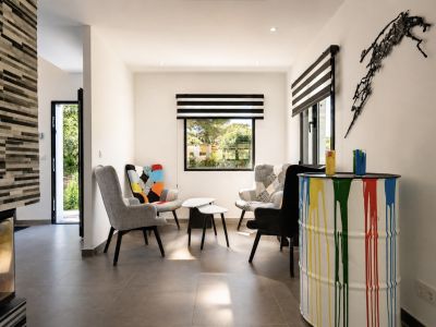 Moderne zithoek met diverse fauteuils, een gestructureerde stenen open haard en een raam met zwart-wit gestreepte jaloezieën. Een kleurrijke verf-druppel ton fungeert als unieke bijzettafel.
