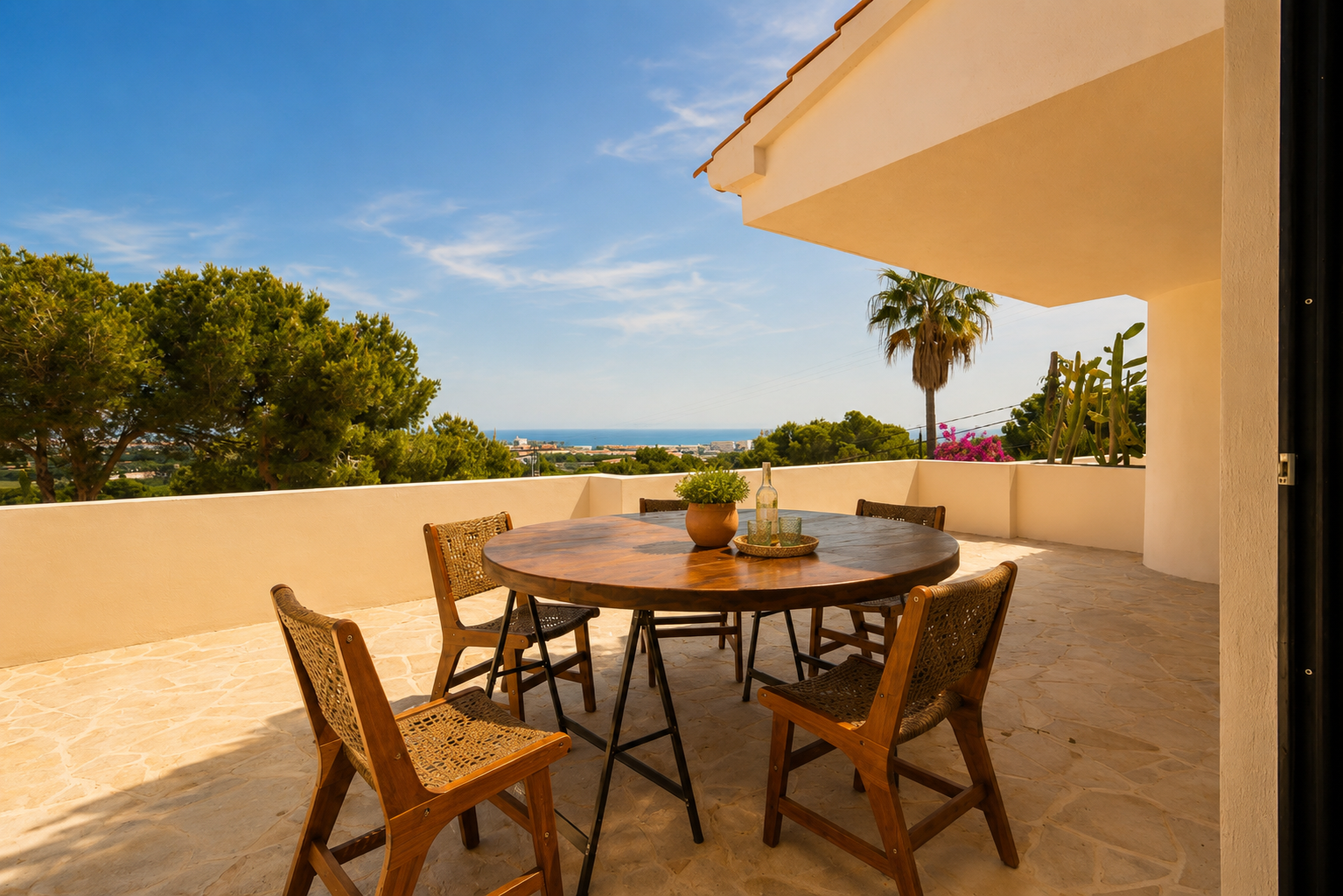 Buitenterras met ronde houten tafel en gevlochten stoelen op plavuizen, met panoramisch uitzicht op de zee en de kustlijn.