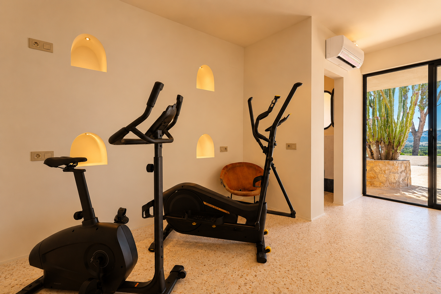Fitnessruimte met hometrainer en crosstrainer, voorzien van verlichte boogvormige wandnissen en uitzicht op een cactustuin en bergen.
