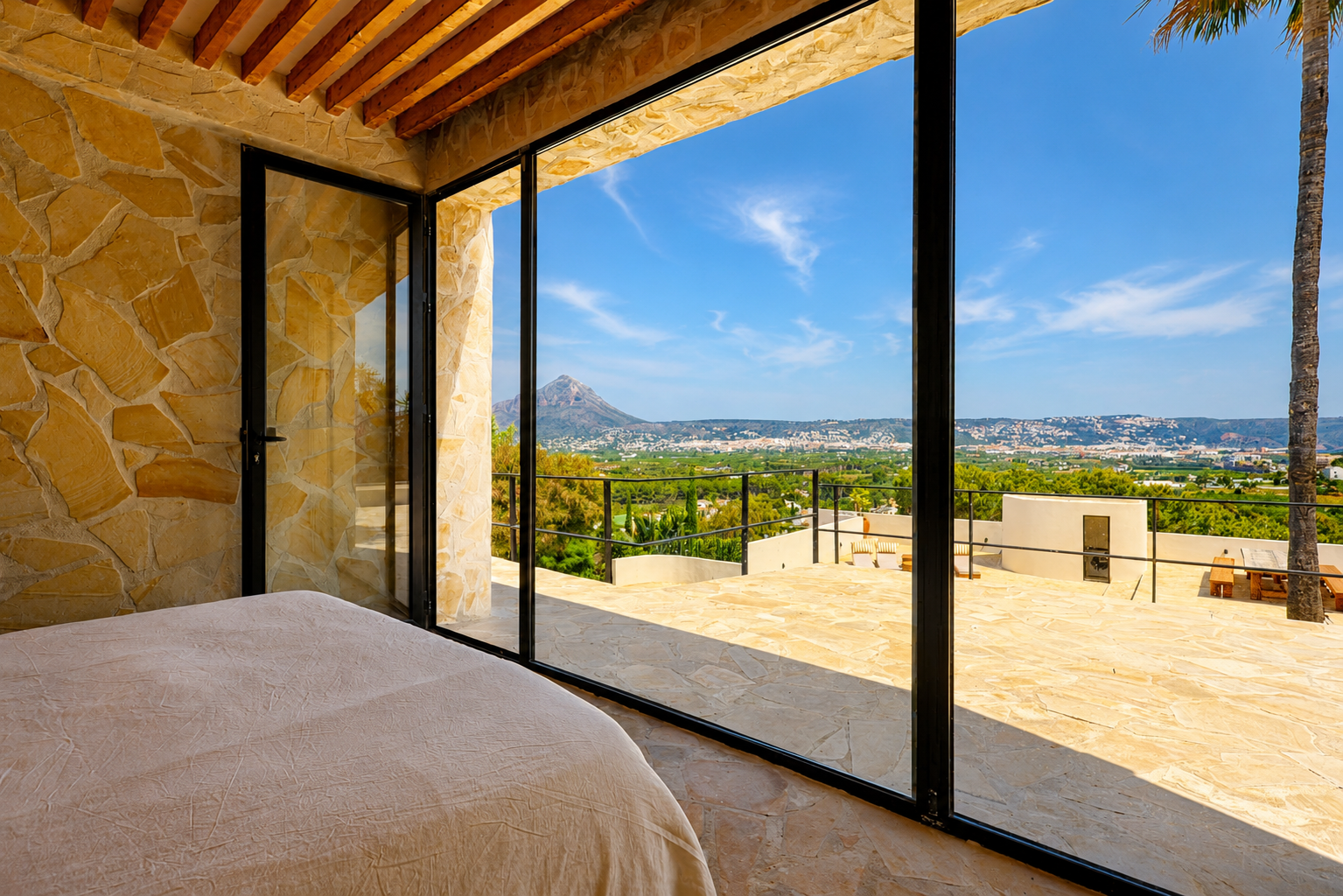Slaapkamer met natuurstenen muren en houten balken, met directe toegang tot een terras met panoramisch uitzicht op de Montgó en de baai van Jávea.
