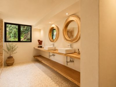 Badkamer met dubbele houten wastafel, twee witte rechthoekige waskommen en grote ronde spiegels met natuurlijke houten lijsten. Een raam biedt uitzicht op groen, passend bij de lichte terrazzovloer.