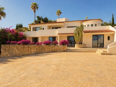 De crèmekleurige gevel van de villa, terracottadakpannen en grote glazen deuren, omzoomd door levendige bougainvillea, een hoge cactus en palmbomen onder een blauwe lucht. Een oprit met patroon leidt naar de ingang.