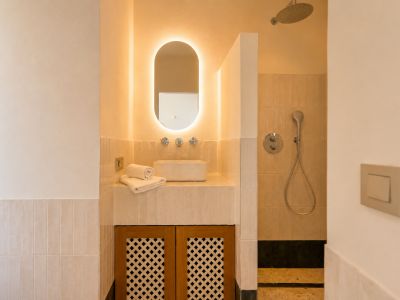 Deze elegante badkamer heeft een opbouw wastafel, een ovale spiegel met achtergrondverlichting en een houten kast met roosterdeuren. De douche heeft een regendouchekop.