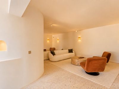 Hedendaagse loungeruimte met gebogen crèmekleurige bank, twee roestkleurige fauteuils en rustieke houten salontafel op lichte terrazzovloer. Geweven wandlampen zorgen voor warme sfeerverlichting.