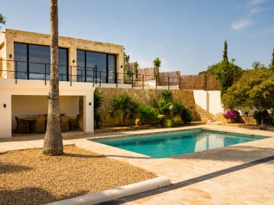 Het terrasvormige exterieur van de villa omvat een rechthoekig zwembad met onregelmatige stenen bestrating, een overdekte eethoek en een stenen gevel met grote ramen.