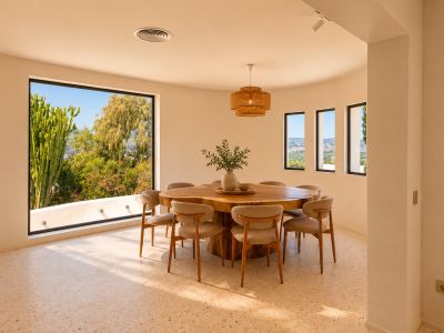Lichte eetruimte met organische houten tafel voor acht personen en terrazzovloer. Grote gebogen ramen bieden uitzicht op weelderig groen en het verre landschap.
