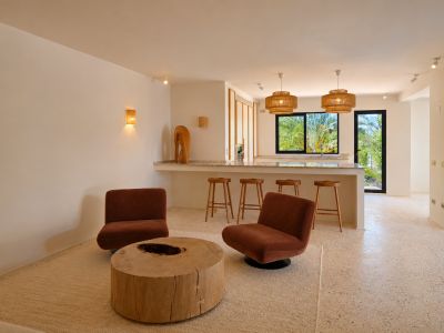Open woonruimte met twee roestkleurige fauteuils, een rustieke houten salontafel en een lichte terrazzovloer. De bar is voorzien van geweven hanglampen en biedt uitzicht op weelderig groen.