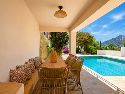 Overdekt buitenterras met houten eettafel en rieten stoelen, direct aan het zwembad. Biedt uitzicht op de Montgó en mediterrane beplanting op een stenen vloer.