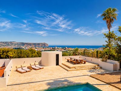 Ruim stenen terras met privézwembad, ligbedden en verhoogde houten eethoek, met panoramisch uitzicht op de baai van Jávea en de kust onder een heldere hemel.