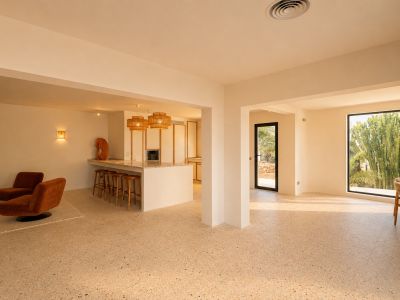 Ruime open woon- en eetkamer met terrazzovloer, inclusief een keukeneiland met bar en een zithoek, verlicht door natuurlijk licht met uitzicht op de weelderige buitenbeplanting.