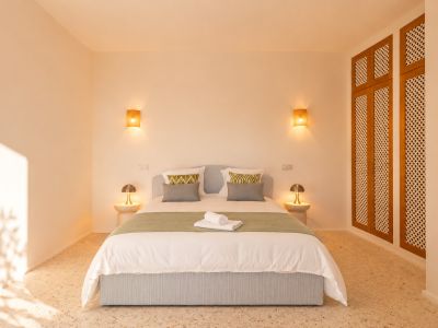 Slaapkamer met lichtblauw gestoffeerd bed, terrazzovloer en warme verlichting van geweven wandlampen. Een houten kast met rasterpanelen is rechts zichtbaar.