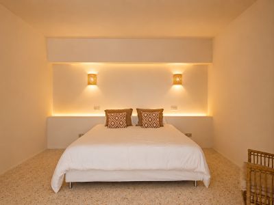 Slaapkamer met tweepersoonsbed, geïntegreerde LED-hoofdbordverlichting en geweven wandlampen. Terrazzovloer en decoratieve kussens.