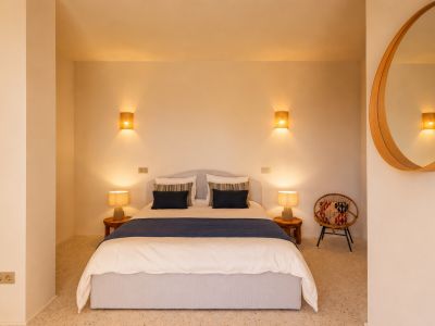 Slaapkamer met tweepersoonsbed, houten nachtkastjes en keramische lampen. Wandlampen met raffia kappen en een ronde houten spiegel sieren de ruimte. Terrazzovloer.