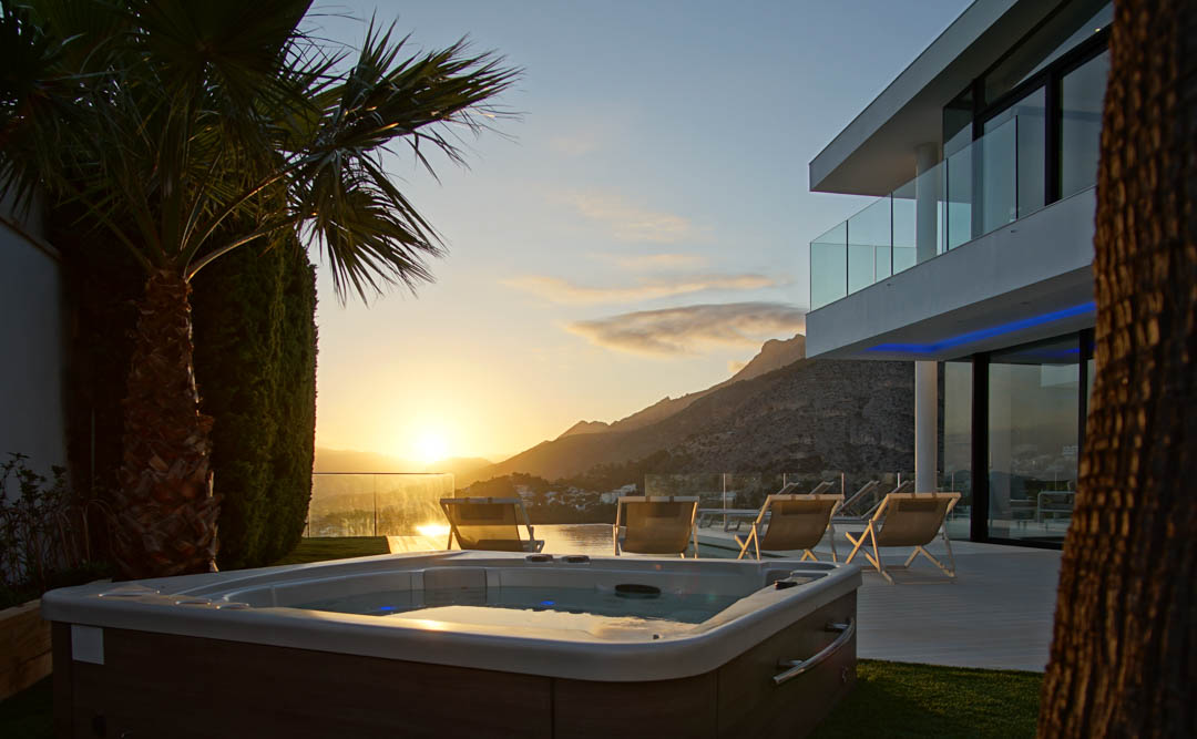 Buitenjacuzzi met houtlook, op groen gras, voor een moderne villa met glazen balkons. Uitzicht op bergen en zee tijdens zonsondergang.