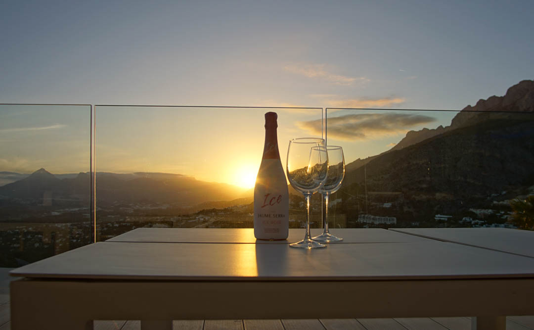 Buitentafel op terras met glazen balustrade, met fles rosé cava en twee glazen. Zonsondergang over bergen en vallei, met warme reflecties van het avondlicht.