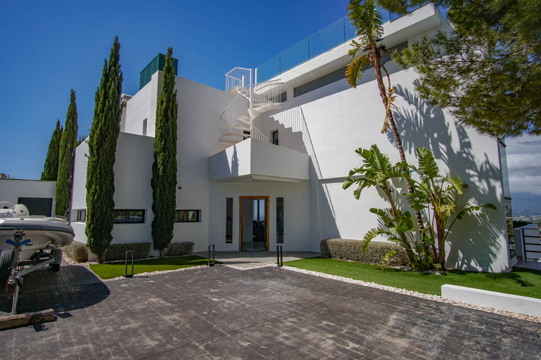 De moderne witte villa-exterieur omvat een geplaveide oprit, hoge cipressen en een opvallende wenteltrap naar het boventerras met glazen balustrade.