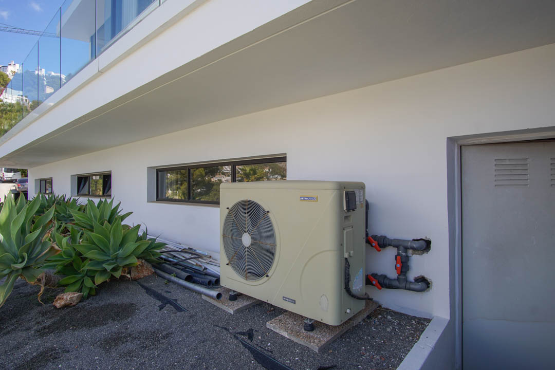 De technische buitenruimte van de villa omvat een AstralPool inverter warmtepomp, horizontale ramen en een technische deur. Agaveplanten sieren de moderne witte gevel.