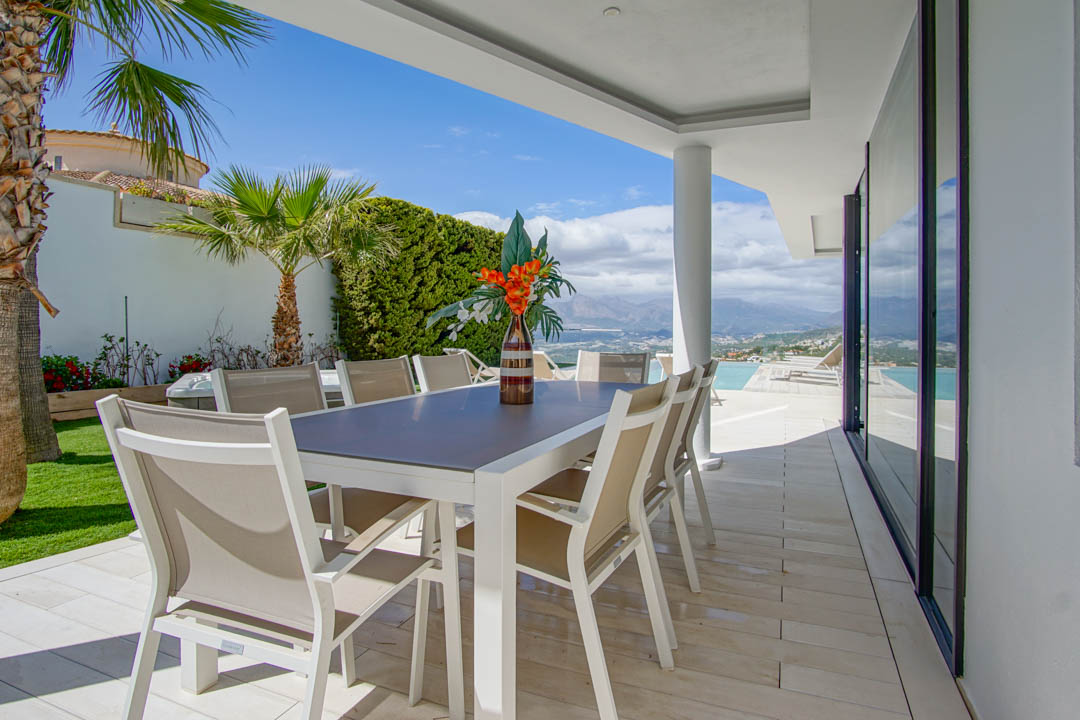 Dit overdekte buitenterras heeft een moderne witte tafel met acht beige stoelen op lichte tegels. Het biedt een weids uitzicht op de bergen en de zee, grenzend aan het overloopzwembad.