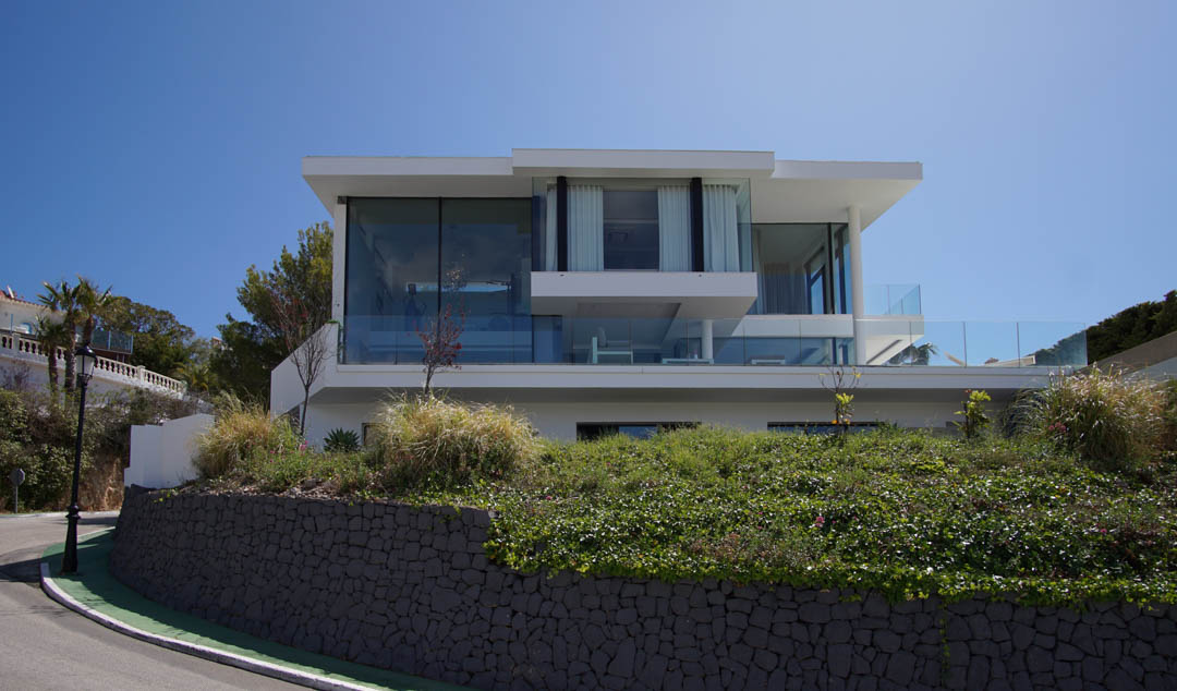 Exterieur van een moderne villa met drie verdiepingen, voorzien van uitgebreide glazen gevels en balustrades, verheven boven een stenen muur en vegetatie langs een bochtige weg.