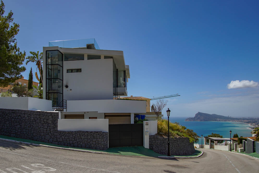 Exterieur van een moderne witte villa met glazen lift en dakterrasreling. Stenen keermuren en een automatische poort omlijsten het panoramische uitzicht op zee.