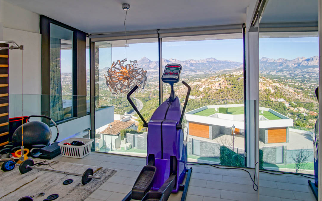 Fitnessruimte met kamerhoge ramen, met panoramisch uitzicht op bergen en vallei. Voorzien van een crosstrainer en gewichten, aangevuld met een glazen balustrade.