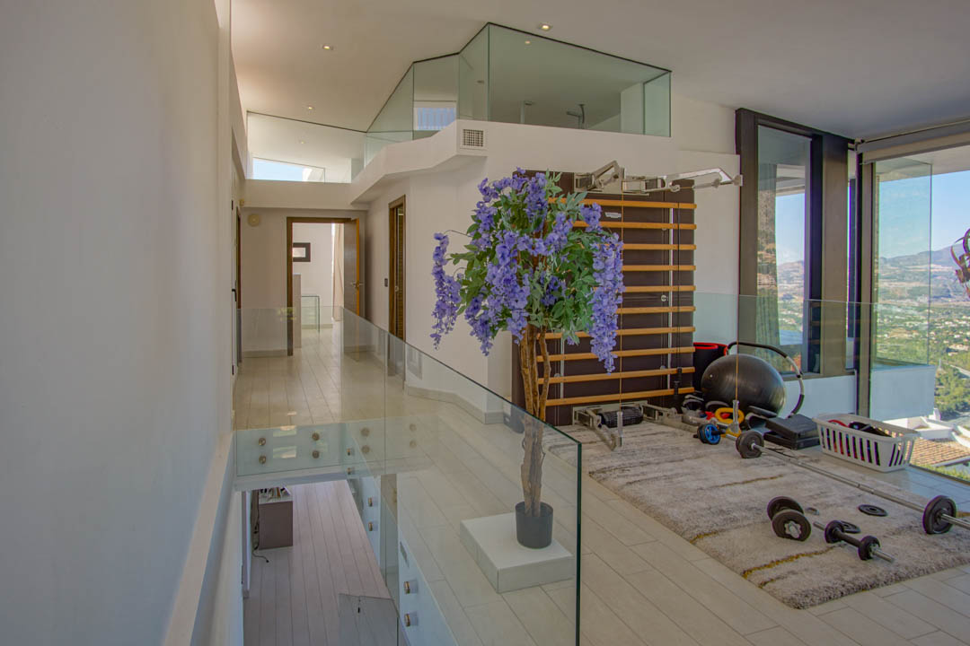 Fitnessruimte op de bovenverdieping met moderne glazen balustrades en lichte vloer. Voorzien van een rekstok en gewichten, biedt het panoramisch uitzicht door kamerhoge ramen.