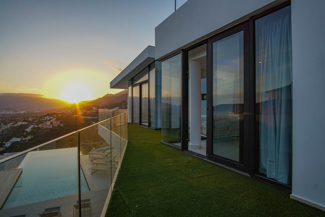 Het ruime terras met overloopzwembad, houten vlonder en glazen balustrade biedt panoramisch uitzicht op de zonsondergang over bergen en vallei.