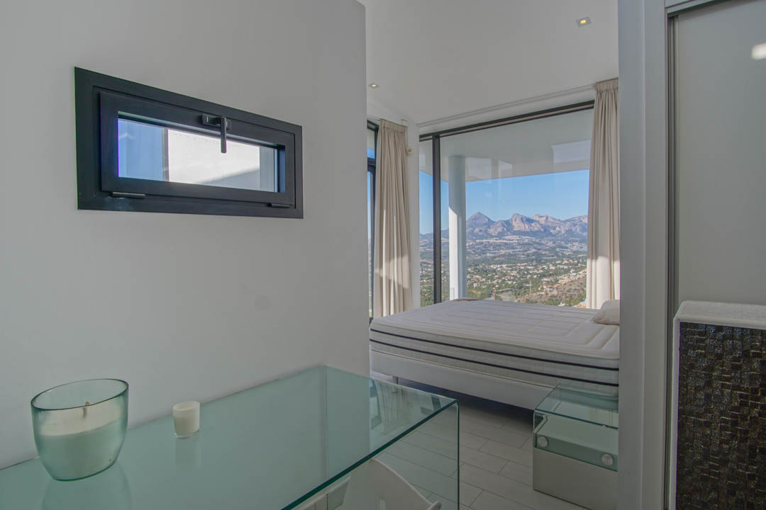 Lichte slaapkamer met bed voor kamerhoge ramen, met panoramisch uitzicht op bergen en vallei. Een glazen console en een horizontaal raam met donker kozijn zijn ook zichtbaar.