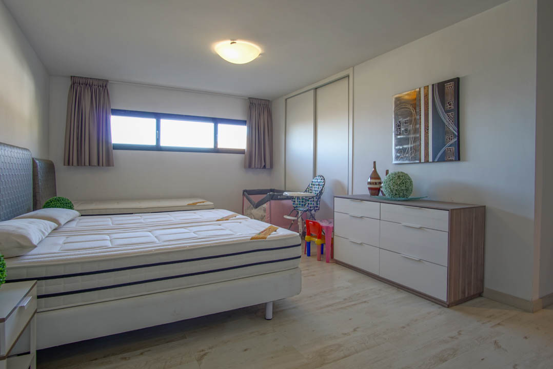 Lichte slaapkamer met een bed van twee matrassen, een lichte houtlook vloer en een langwerpig horizontaal raam. De kamer omvat een witte schuifdeurkast en een moderne ladekast met abstracte kunst erboven.