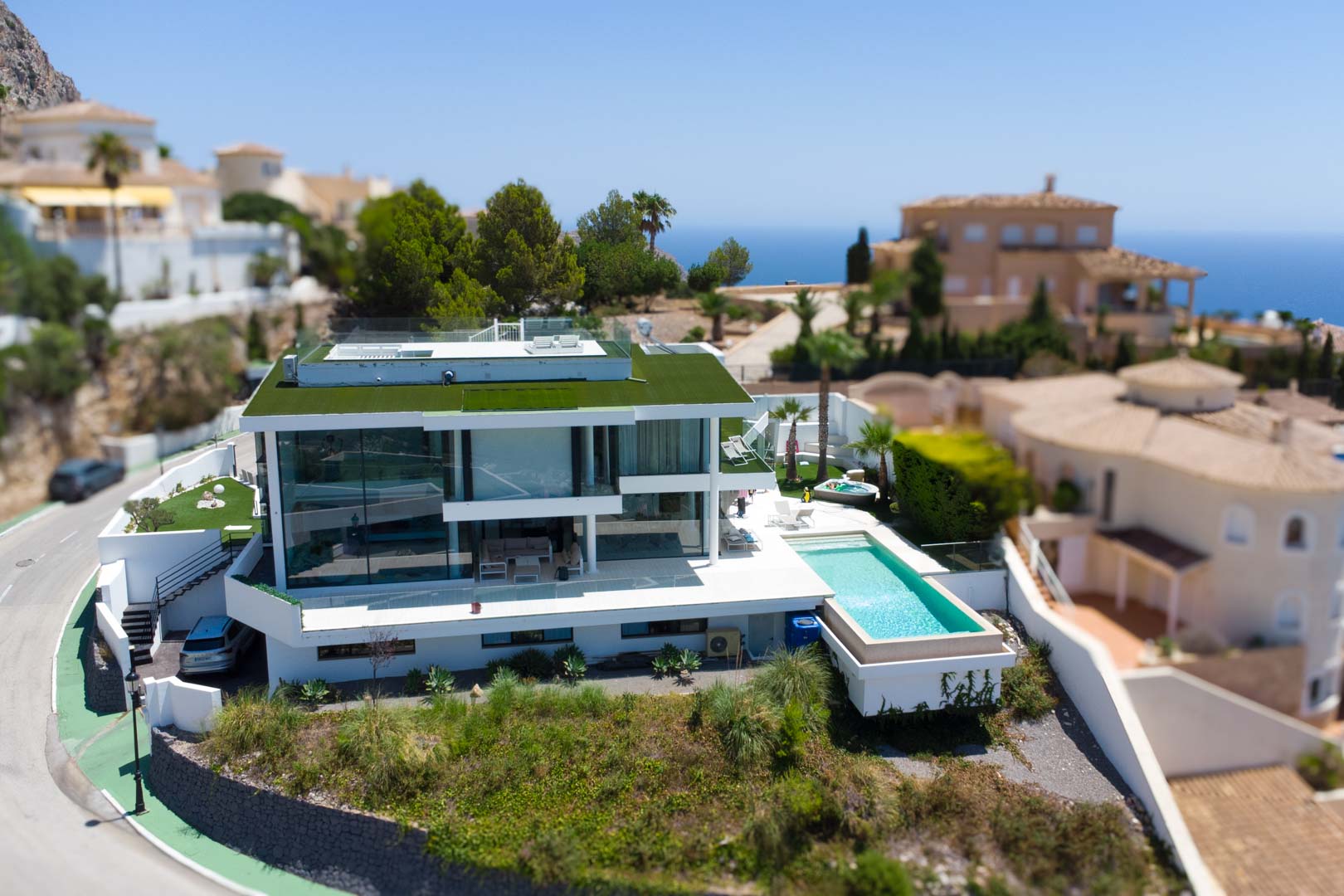 Luchtfoto van de moderne witte designvilla met groen dak, overloopzwembad en aparte jacuzzi, gelegen in een omgeving met zeezicht.