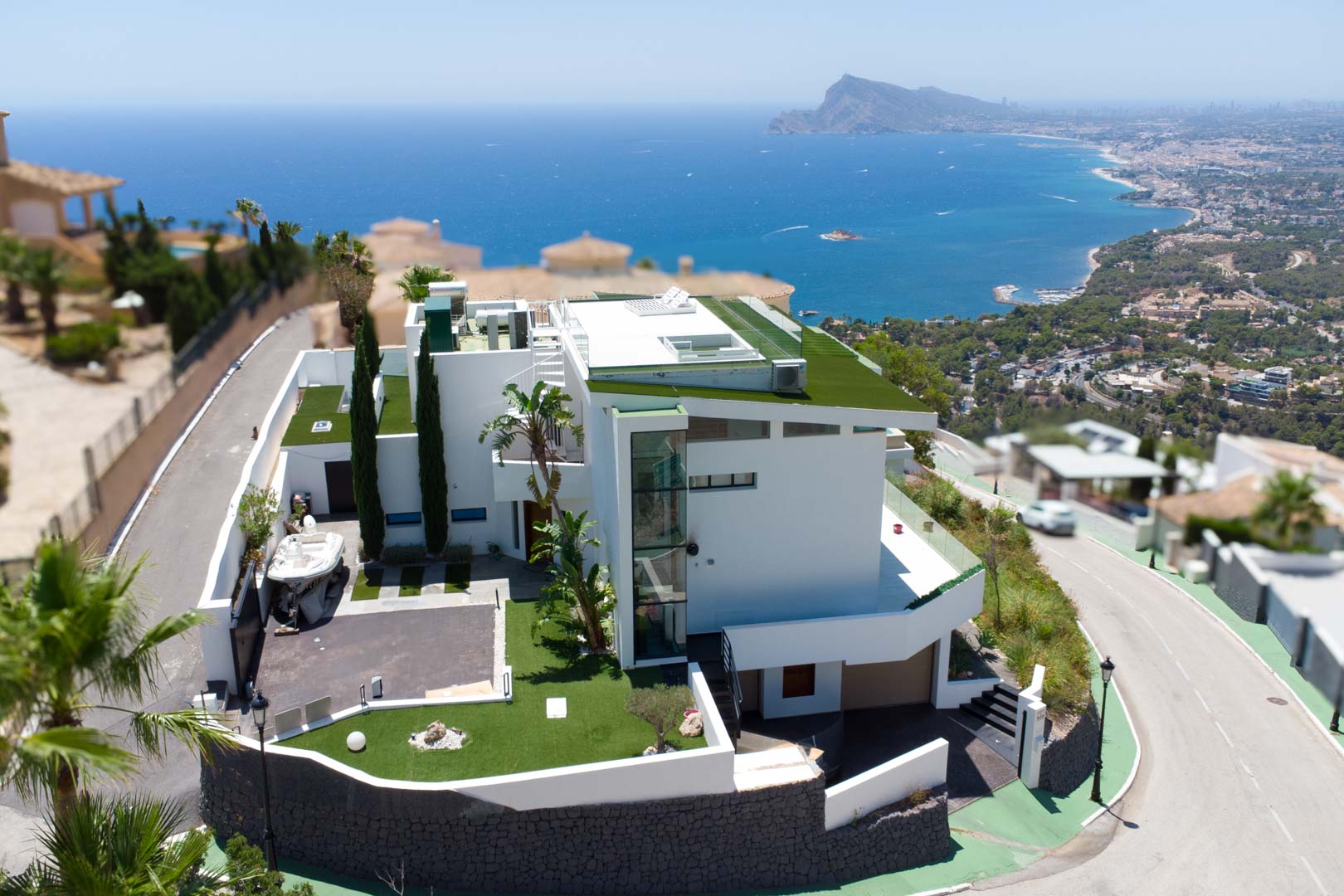 Luchtfoto van een moderne witte villa met architectuur op meerdere niveaus, groene platte daken en een externe glazen lift. Het pand omvat een privé-oprit en biedt panoramisch zee- en kustzicht.