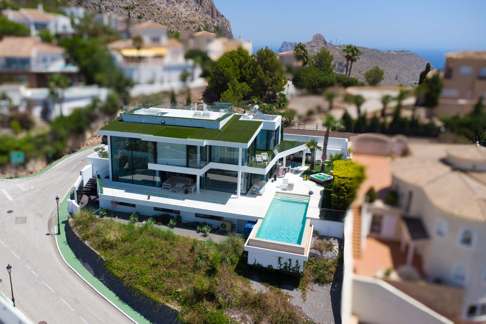 Luchtfoto van een moderne witte villa met overloopzwembad, glazen gevels, een groen dak en aparte jacuzzi, met uitzicht op bergen en zee.