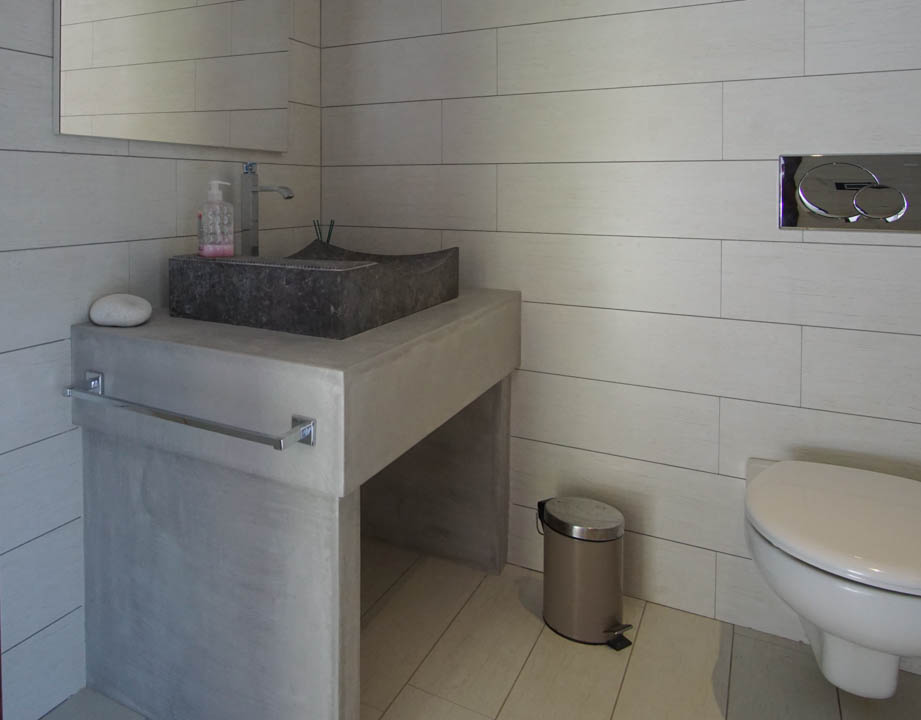 Modern gastentoilet met stenen waskom op betonnen wastafelmeubel en lichte tegels. Wandhangend toilet met chromen spoelplaat.
