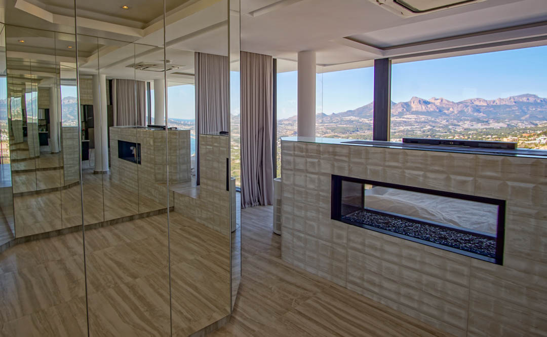 Modern interieur met een transparante gashaard op kiezelbasis, ingebouwd in een getextureerde muur. Spiegelpanelen reflecteren de kamer en het weidse berg- en zeezicht door kamerhoge ramen.