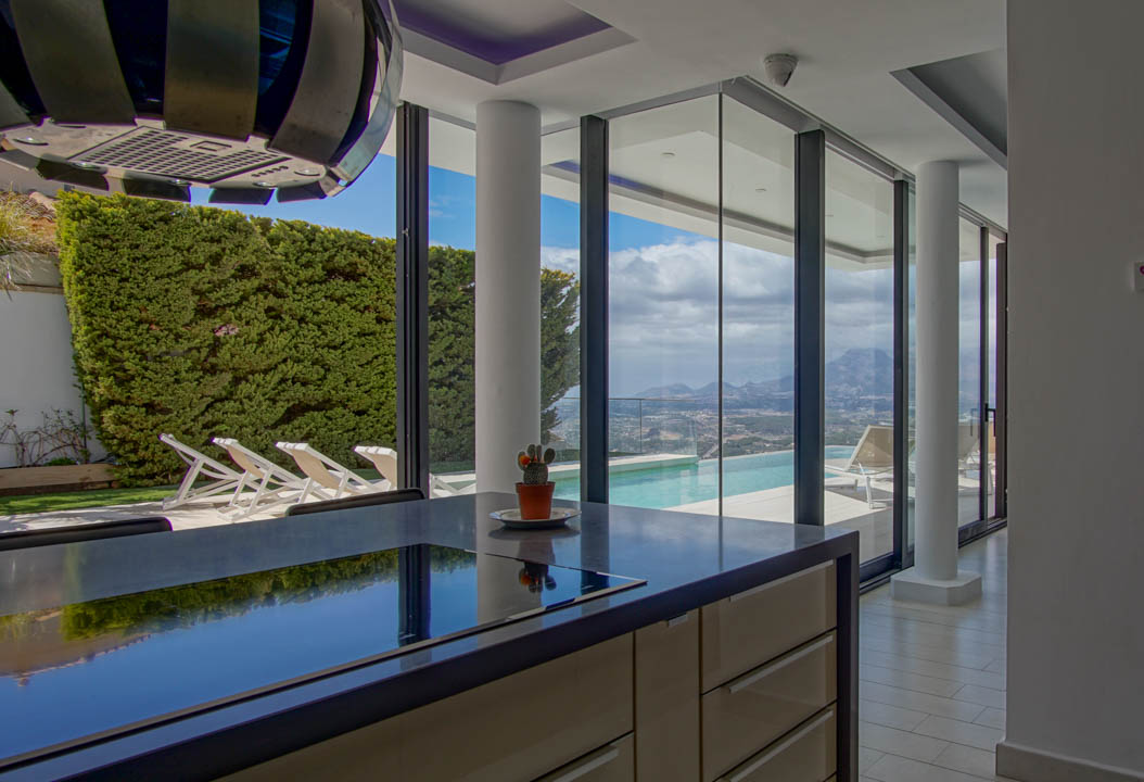 Modern kookeiland met donker, reflecterend werkblad en geïntegreerde kookplaat. Grote glazen deuren bieden uitzicht op de infinity pool en panoramische berg- en dalgezichten.