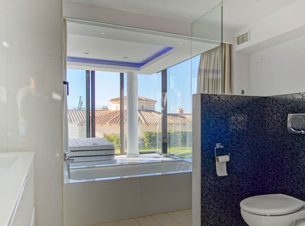 Moderne badkamer met ingebouwd bad en kamerhoge ramen met uitzicht op een pannendak en tuin. Voorzien van blauwe accentverlichting en een donkergepatroneerde scheidingswand.
