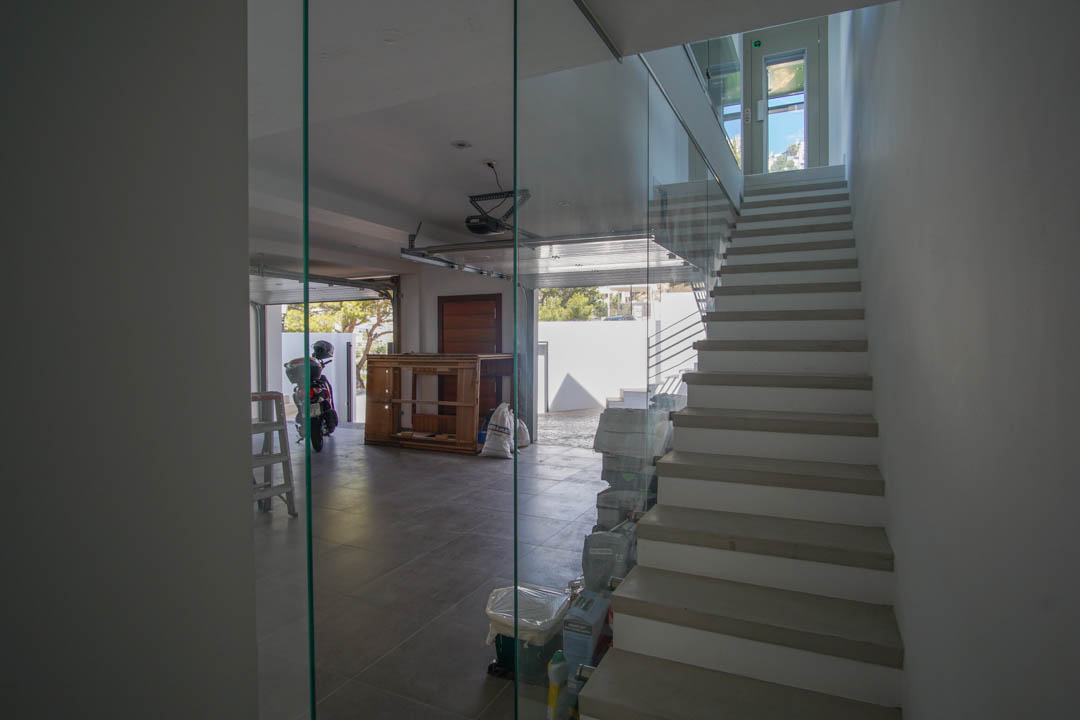 Moderne binnentrap met glazen balustrade en lichte treden, grenzend aan een betegelde garage met open deur en natuurlijk daglicht.
