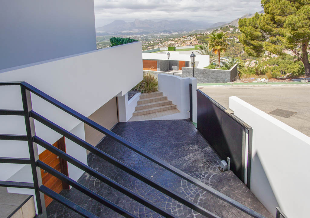 Moderne buitentrap met zwarte balustrades daalt af naar een donkergrijze, gestempelde oprit, voorzien van een automatische poort, garage-ingang en panoramisch bergzicht.
