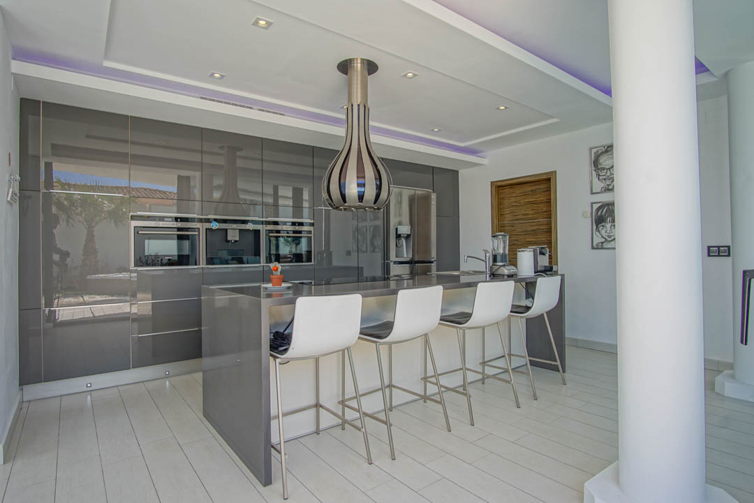 Moderne keuken met hoogglans grijze kasten, geïntegreerde ovens en een centraal eiland met zitplaatsen voor vier. Inbouwspots en paarse LED-stripverlichting in het plafond.