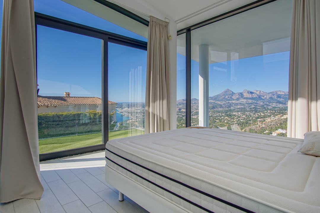 Moderne slaapkamer met groot bed en uitgestrekte ramen. Het biedt panoramisch uitzicht op de verre zee, een groen gazon en ruige bergen onder een heldere hemel.