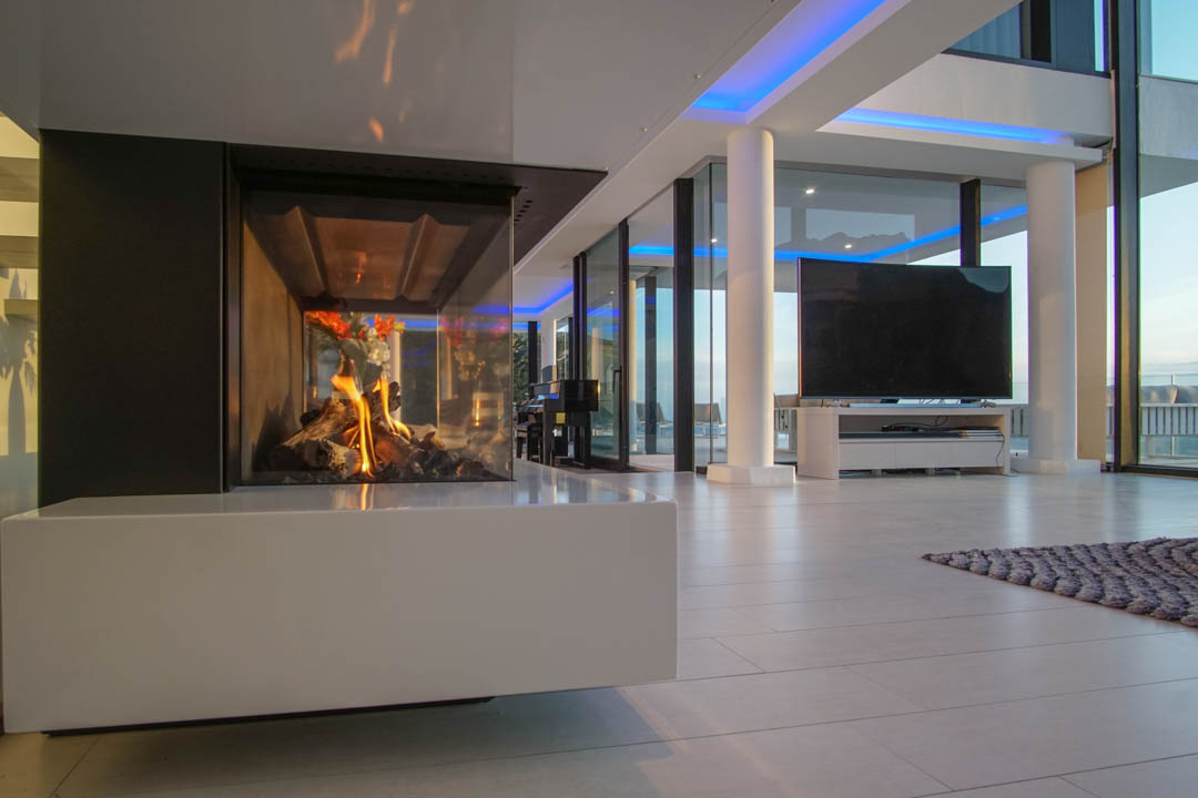 Moderne woonruimte met glazen gashaard en witte basis. Grote ramen en blauwe accentverlichting complementeren het ontwerp.