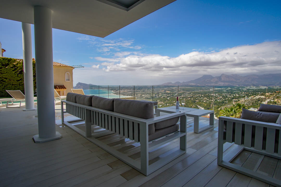 Ruim overdekt terras met lichte vlonder en modern wit loungemeubilair, dat via een glazen balustrade een panoramisch uitzicht op zee, kust en bergen omlijst.