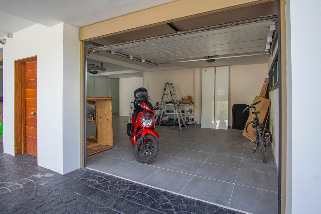 Ruime garage met grijze tegelvloer, een deels geopende sectionaaldeur en een opvallende houten toegangsdeur links. Binnen zijn een rode scooter en fiets zichtbaar.