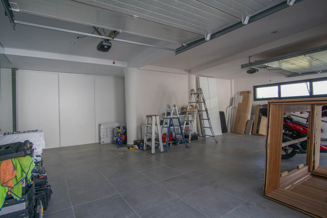 Ruime garage met grijze tegelvloer en witte muren. Voorzien van twee automatische poortmechanismen, horizontale ramen en witte schuifdeuren voor opslag.