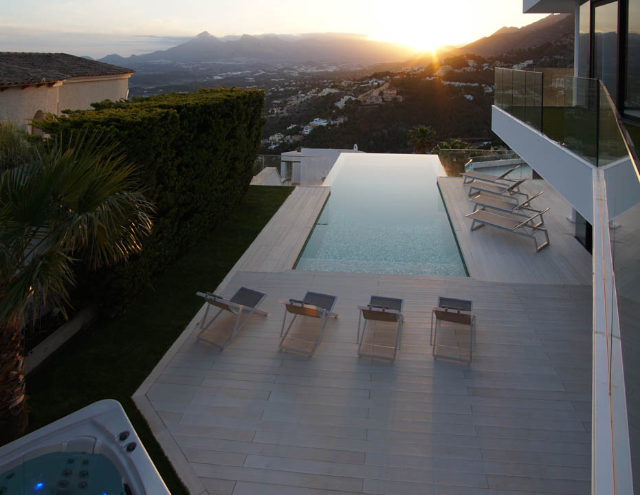 Ruime infinity pool met lichtgekleurd terras, diverse ligbedden en aparte jacuzzi, omkaderd door een panoramisch berglandschap bij zonsondergang.