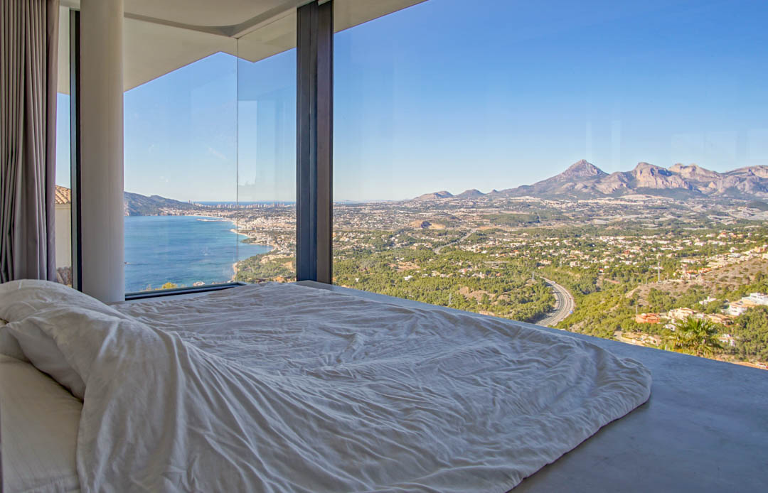 Slaapkamer met kamerhoge glazen ramen, die panoramisch uitzicht bieden op de Middellandse Zee, kustplaats en bergen. Een laag bed met wit beddengoed kijkt uit op het weidse panorama.
