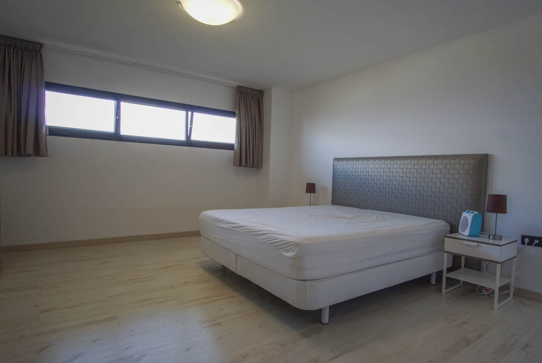 Slaapkamer met lichte houtnerf vloer en witte muren. Voorzien van een bed met gestructureerd hoofdbord, nachtkastje en een langwerpig raam met donkere kozijnen.